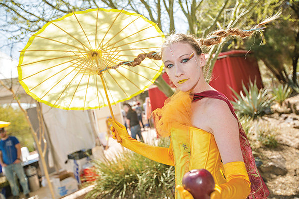 A women in a yellow costume with wacky hair holds a tiny umbrella.